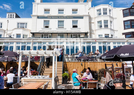Seafront Royal Albion Hotel. Foreground, people sitting at tables in bright sunshine. Behind them, veranda main white hotel building. Blue sky. Stock Photo