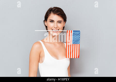 Happy young woman holding USA flag in teeth and winking over gray background Stock Photo