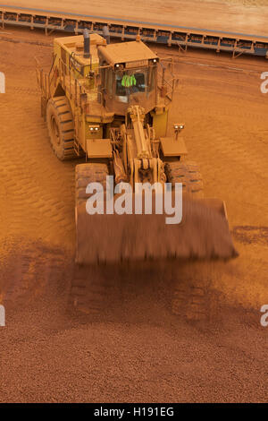 Front end loader loading hopper with lump iron ore from reclaimed stack behind. Ore then passes onto conveyor belt and loaded onto ship in port. Stock Photo