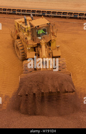 Front end loader loading hopper with lump iron ore from reclaimed stack behind. Ore then passes onto conveyor belt and loaded onto ship in port. Stock Photo