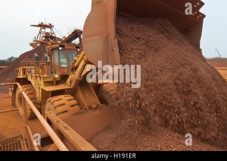 Front end loader loading hopper with lump iron ore from reclaimed stack behind. Ore then passes onto conveyor belt and loaded onto ship in port. Stock Photo