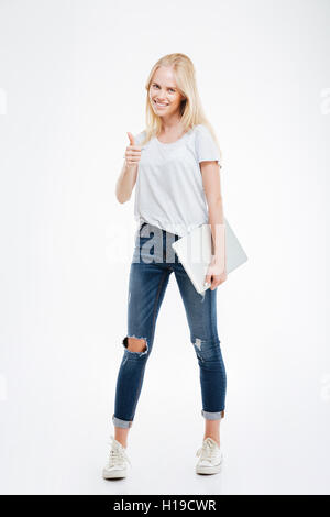 Full length portrait of a beautiful girl holding a laptop and giving thumb up gesture isolated on white Stock Photo
