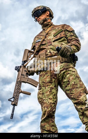 Young handsome military man holding automatic rifle and hand grenade on sky background. Airsoft Stock Photo