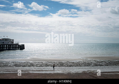 A person walks along the beach in Worthing, West Sussex, England near to Worthing Pier during low tide on a sunny day. Stock Photo