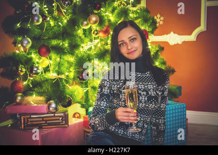 Beautiful brunette girl with a glass of champagne and a luxurious smile near Christmas tree in room Stock Photo