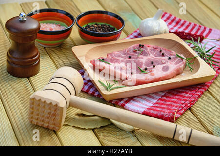 Raw pork meat on wooden plate, pepper mill, rosemary, spices and hammer on wooden background Stock Photo