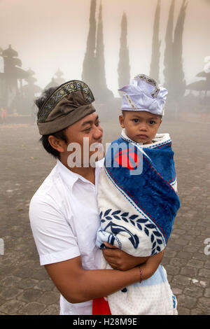 Indonesia, Bali, Batur, Pura Ulun Danu Batur, man wearing traditional udeng hat carrying young son wrapped in blanket Stock Photo