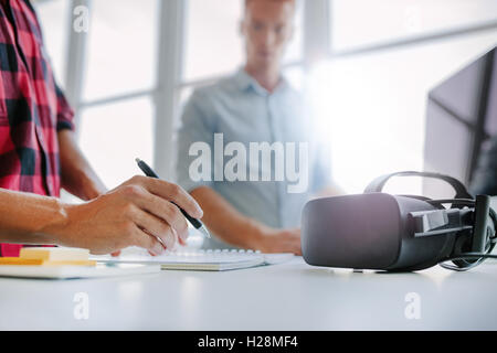 Virtual reality glasses on table with two men working in office.  Man writing colleague using computer in background. Stock Photo