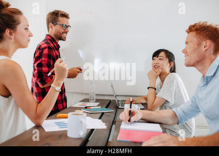 Group of young people sitting around conference table with male colleague standing by white board for presentation. Startup asso Stock Photo