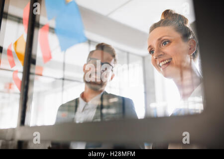 Portrait of two young professionals looking over adhesive notes on glass wall and brainstorming. Young man and women standing at Stock Photo