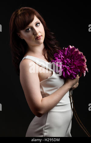 Auburn brunette english rose female portrait, black backdrop, purple lighting, wearing white jumpsuit with purple flower Stock Photo