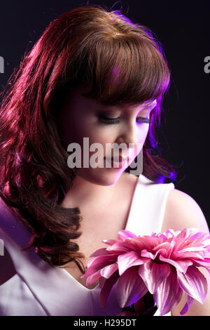 Auburn brunette english rose female portrait, black backdrop, purple lighting, wearing white jumpsuit with pink flower Stock Photo