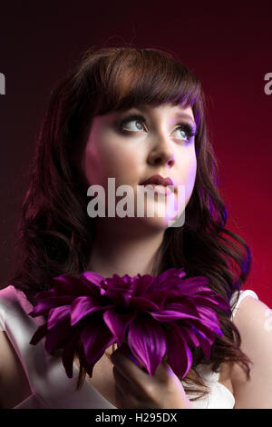 Auburn brunette english rose female portrait, red backdrop, purple lighting, wearing white jumpsuit with purple flower Stock Photo