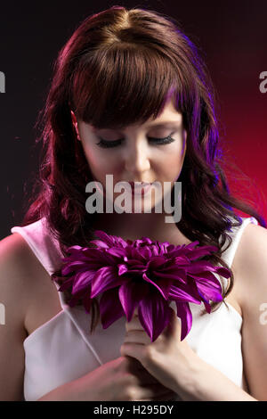 Auburn brunette english rose female portrait, red backdrop, purple lighting, wearing white jumpsuit with purple flower Stock Photo