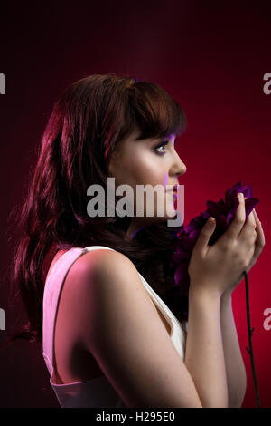 Auburn brunette english rose female portrait, red backdrop, purple lighting, wearing white jumpsuit with purple flower Stock Photo