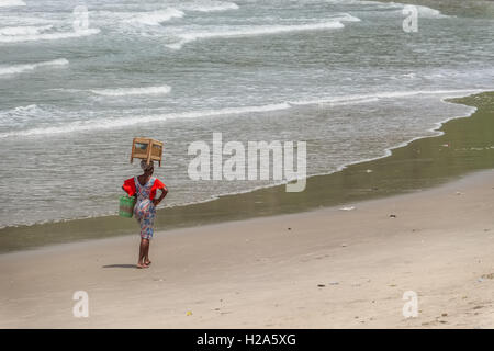 Female beach seller with cabinet of food on top of her head walking along beach in Abandze Beach in Ghana Stock Photo