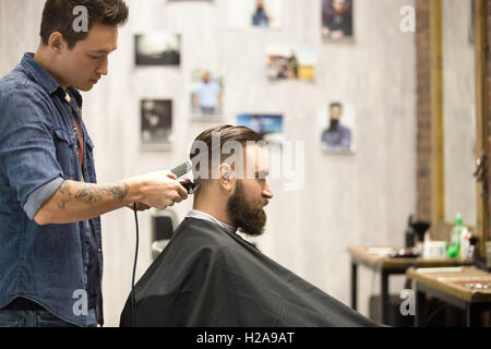 Side view portrait of handsome young bearded caucasian man getting trendy haircut in modern barbershop. Cool men hair stylist wi Stock Photo