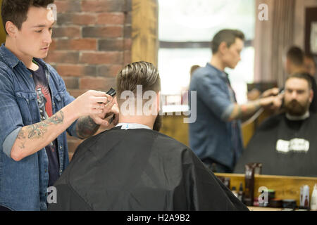 Back view of handsome young bearded man getting trendy haircut in modern barbershop. Cool male hairstylist with tattoo "born bar Stock Photo