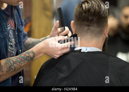 Close-up back view of handsome young bearded man getting trendy haircut in modern barbershop. Cool male hairstylist with tattoo Stock Photo