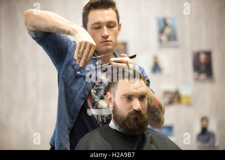 Portrait of handsome young bearded man getting trendy haircut in modern barbershop. Cool male hairstylist with tattoo serving cl Stock Photo