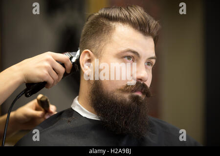 Side view portrait of handsome young bearded caucasian man getting trendy haircut in modern barbershop. Attractive barber girl w Stock Photo
