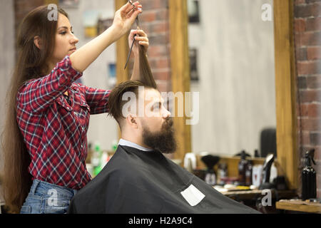 Side view portrait of handsome young bearded caucasian man getting trendy haircut in modern barber shop. Attractive hairstylist Stock Photo