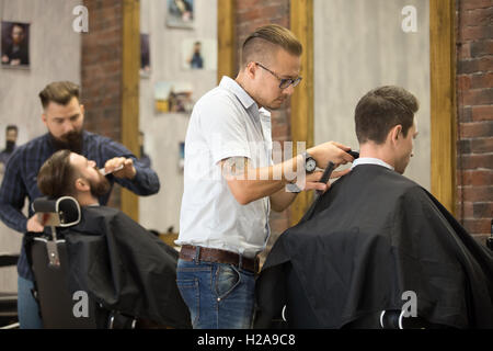 Interior shot of working process in barbershop. Back view of handsome young men getting trendy haircuts in modern barbershop. Co Stock Photo