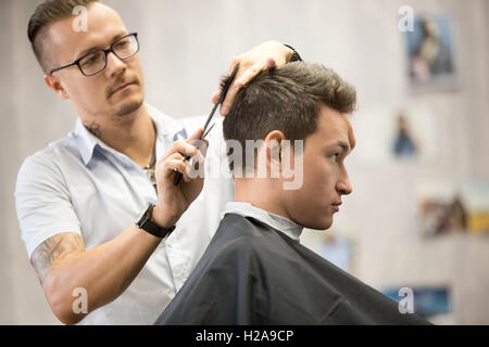 Interior shot of working process in modern barbershop. Side view portrait of attractive young man getting trendy haircut. Male h Stock Photo