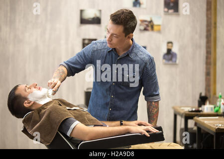 Interior shot of working process in modern barbershop. Side view portrait of handsome young man getting beard shaving. Male hair Stock Photo