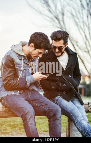 Two trendy casual young men sitting together on a bench outdoors reading an sms or text message, or looking at photos on a mobil Stock Photo