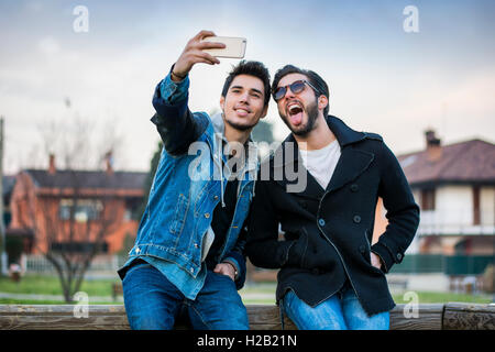 Two young men taking selfie while sitting on bench outdoors Stock Photo