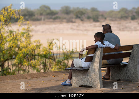 Father and son sitting on a bench in the shade enjoying the Letaba River Stock Photo