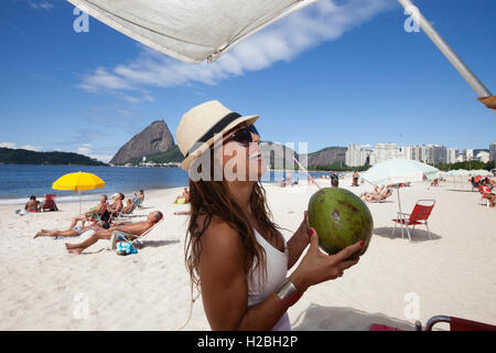 Pretty brunette Brazilian woman drinks coconut water at Flamengo beach, Sugarloaf mountain in background, Rio de Janeiro, Brazil. Stock Photo