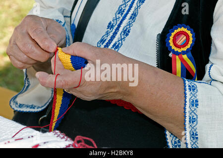 Old woman dressed in romanian traditional clothing, interweaves by hand one belt in national colors and design. Stock Photo