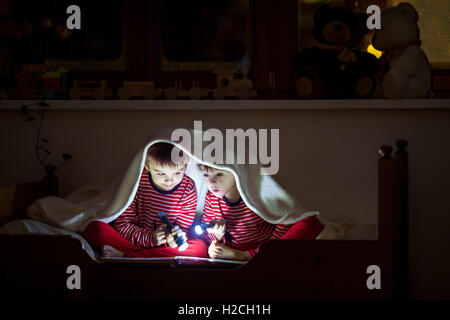 Two sweet boys, reading a book in bed after bedtime, using flashlights, winter night Stock Photo