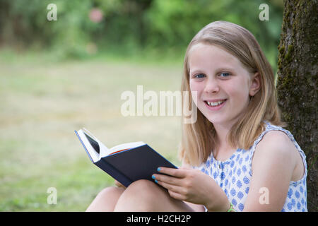 Girl Sitting Against Tree In Garden Reading Book Stock Photo