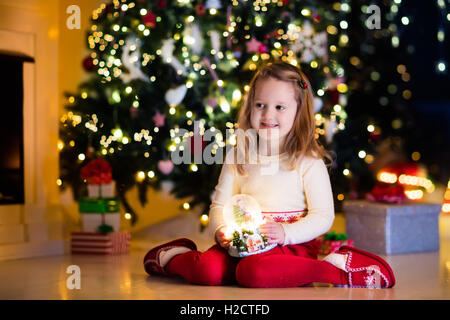 Family on Christmas eve at fireplace. Little girl opening Xmas presents holding snow globe. Child under Christmas tree Stock Photo