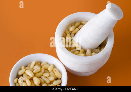 Shelled pine nuts in white bowl and mortar on orange background. Edible seeds of Korean pines family Pinaceae, Pinus koraiensis. Stock Photo