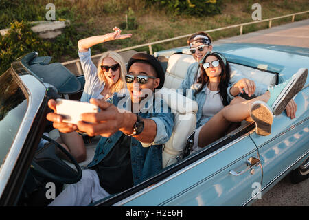 Multiethnic group of joyful young friends taking selfie with mobile phone in cabriolet Stock Photo