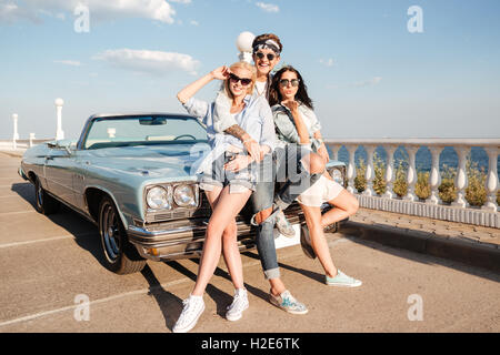 Full length of cheerful young man and two happy women standing near vintage cabriolet in summer Stock Photo