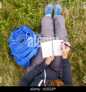 Woman with blue backpack sitting on grass and writing into blank notebook. Overhead view