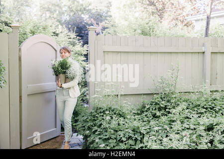 Woman entering a garden through a gate, carrying a bunch of white flowers. Stock Photo