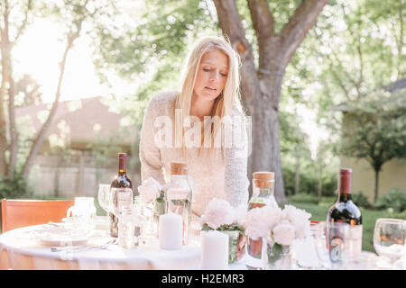 blonde woman setting a table in a garden, vases with pink roses. Stock Photo
