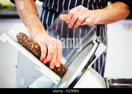 Close up of a butcher wearing a striped blue apron , slicing salami with a slicer. Stock Photo