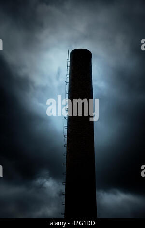 Dark and ominous sky and silhouette of factory chimney. Heavy clouds and darkness. Moody and scary setting in industrial area. Stock Photo