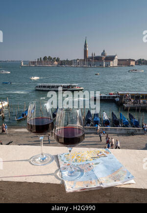 Two glasses of red wine and tourist map on hotel balcony overlooking moored gondolas and church of S. Giorgio Maggiore.  Venice Stock Photo