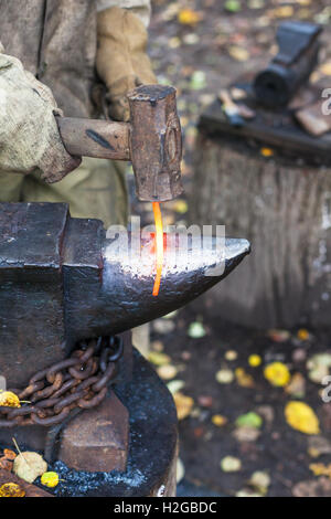 Blacksmith forges red hot glowing iron rod on anvil in outdoor rural smithy Stock Photo