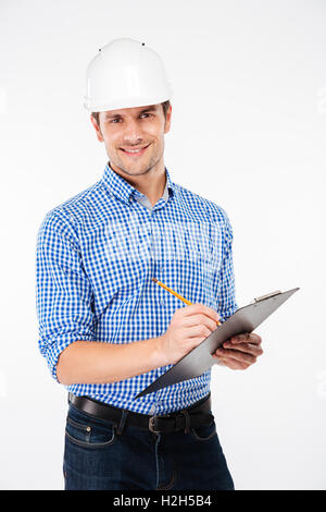Smiling young man architect in building helmet standing and writing on clipboard Stock Photo