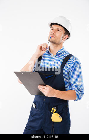 Pensive young builder in helmet thinking and writing on clipboard isolated on a white background Stock Photo