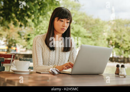 Shot of attractive young woman sitting at cafe table outdoors and working on laptop. Asian female surfing internet on laptop com Stock Photo
